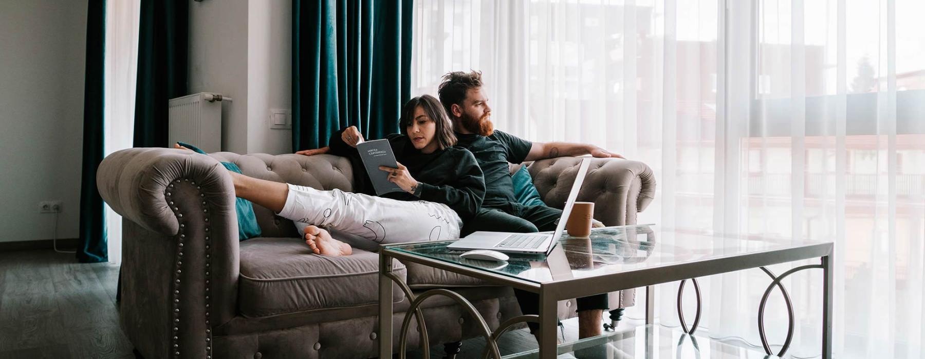 man sits on a couch and looks out the window while his wife lies against him reading a book